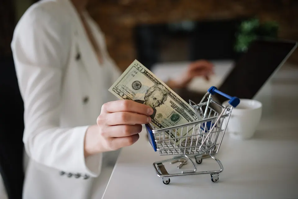 A woman at a desk shopping online with a small toy shopping cart and cash beside the laptop
